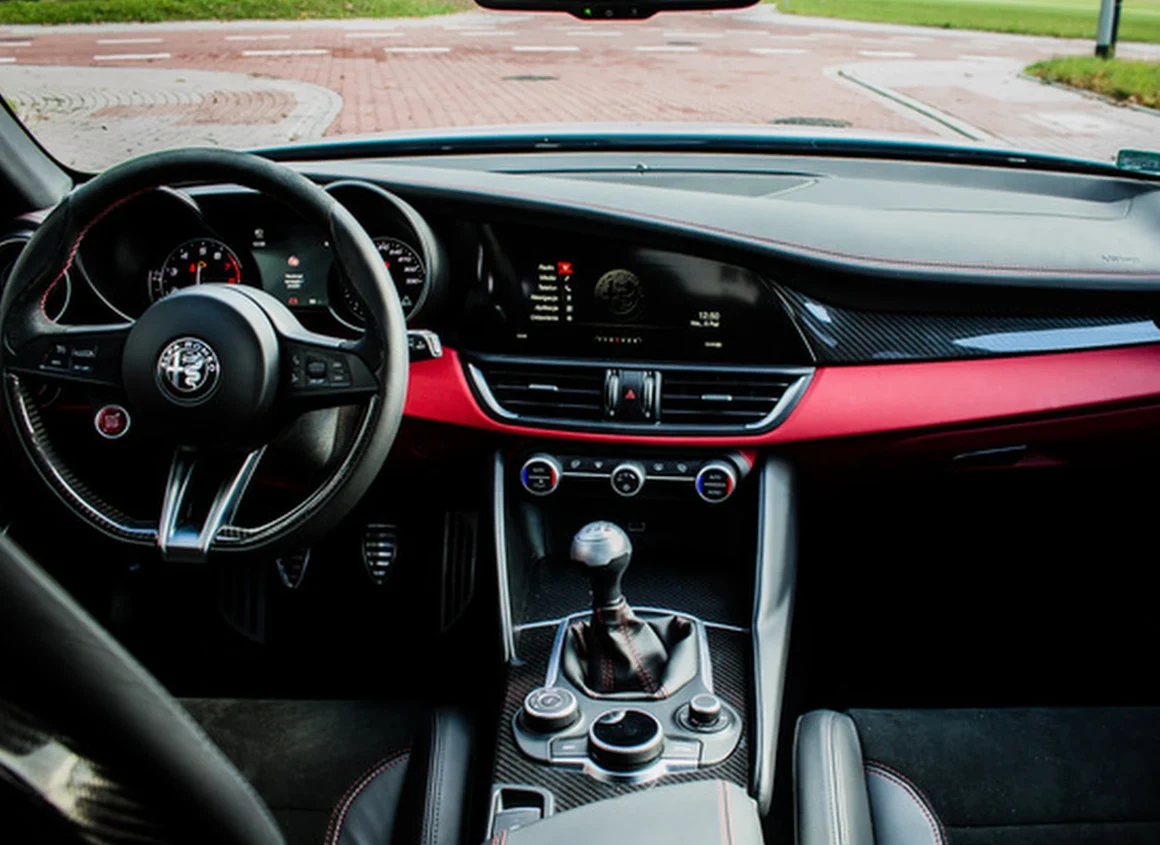 wnętrze samochodu Alfa Romeo z widocznym sportowym kierownicą, manualną skrzynią biegów, elektronicznym wyświetlaczem na desce rozdzielczej oraz czerwonym elementem dekoracyjnym biegnącym przez całą deskę