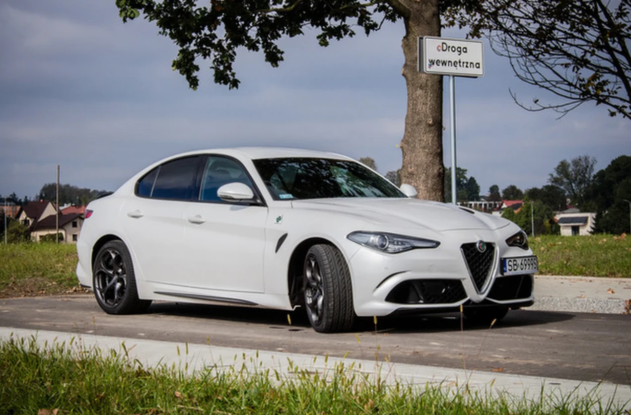 biały samochód Alfa Romeo Giulia zaparkowany na asfaltowej drodze przy krawężniku, na tle pojedynczego drzewa i tabliczki z napisem „Droga wewnętrzna”