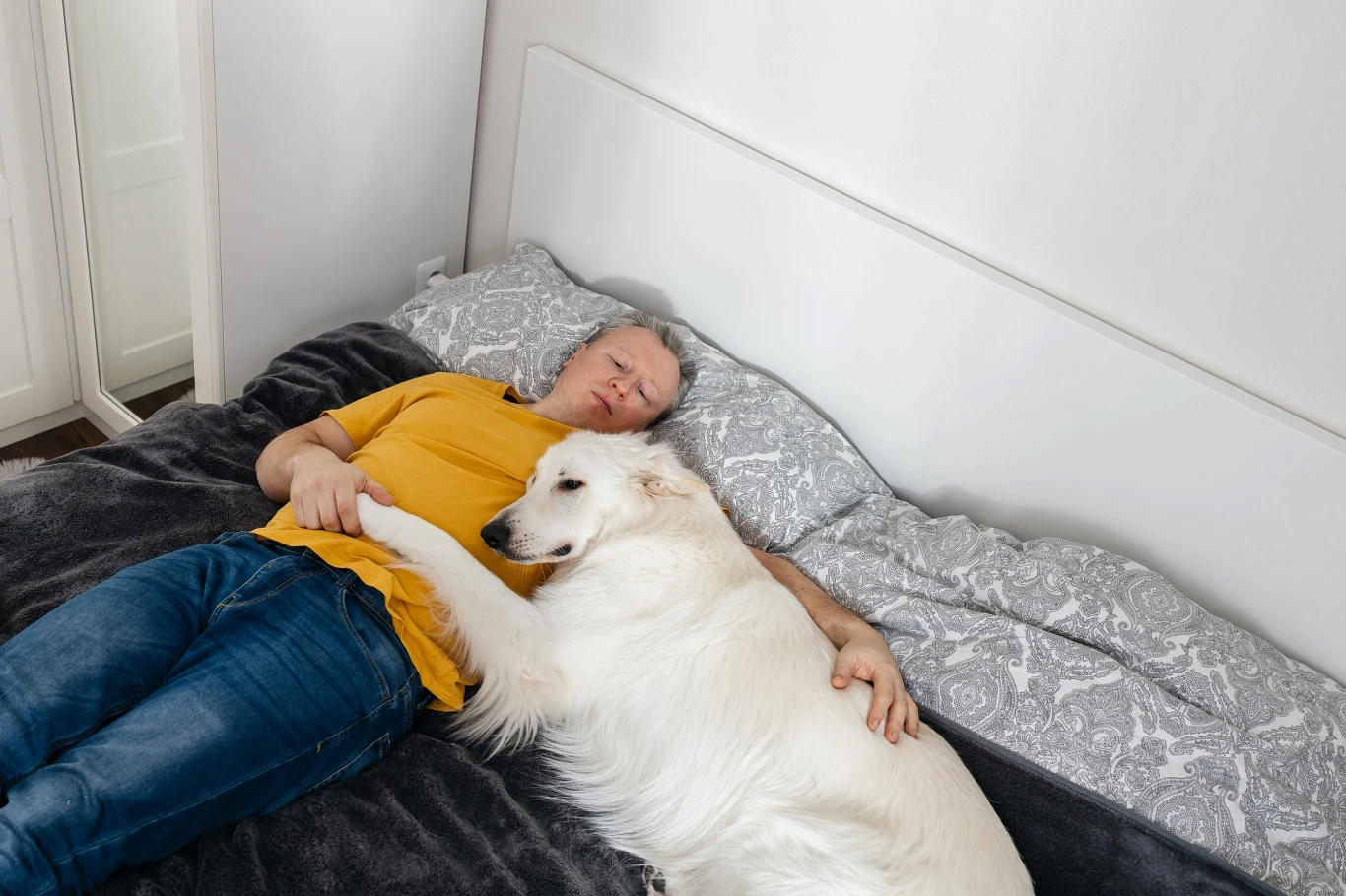 Mężczyzna w żółtej koszulce leży na łóżku, przytulając dużego, białego psa wtulonego w jego ramię.