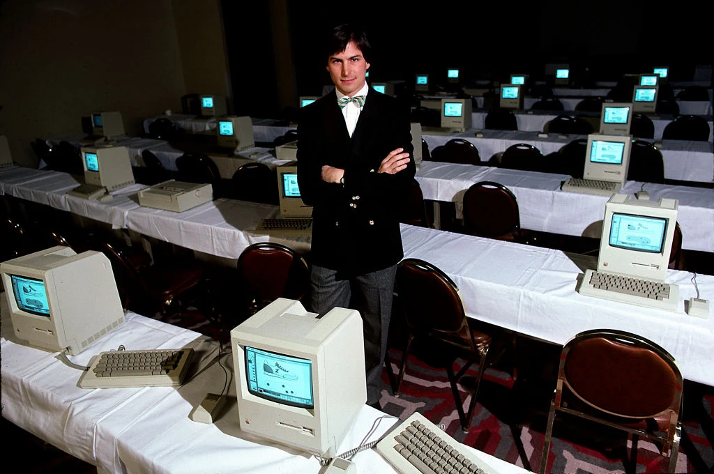 mężczyzna w garniturze stoi w sali pełnej komputerów Macintosh ustawionych w równych rzędach na białych stołach, wszystkie monitory wyświetlają ten sam ekran, pomieszczenie o półmrocznym oświetleniu