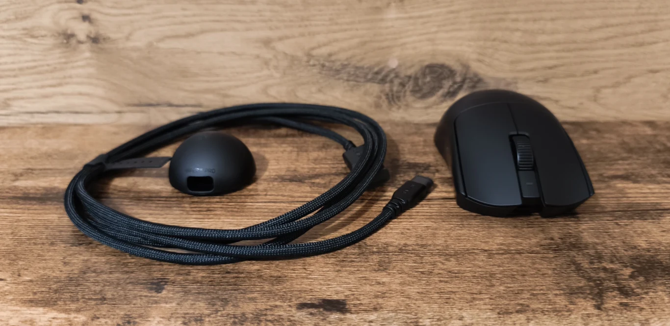 Czarna mysz komputerowa obok odłączanego przewodu USB w oplocie materiałowym owiniętego wokół czarnego adaptera, wszystko na drewnianym blacie z jasnym tłem.