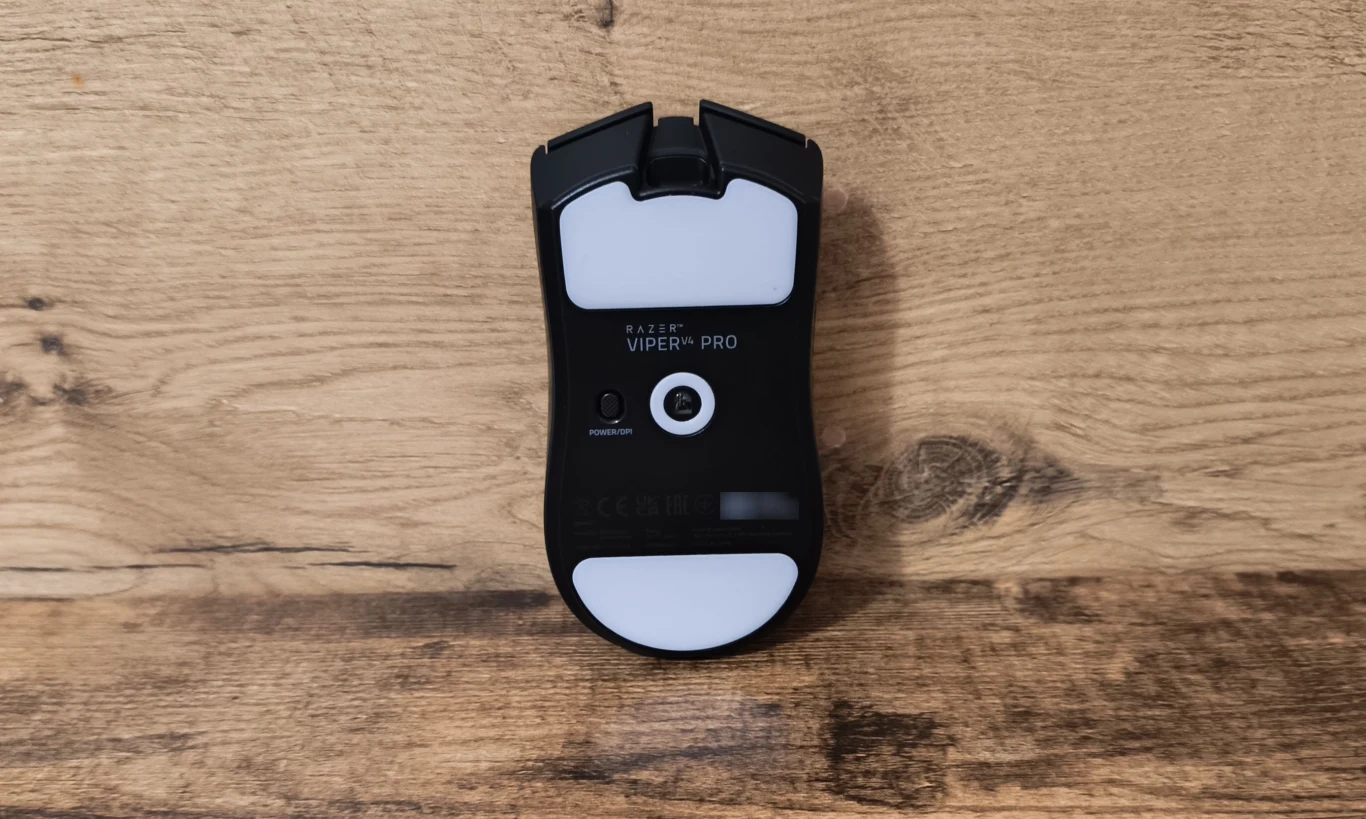 czarna myszka komputerowa Razer Viper V2 Pro położona spodem do góry na drewnianej powierzchni, widoczne duże białe ślizgacze oraz sensor optyczny pośrodku