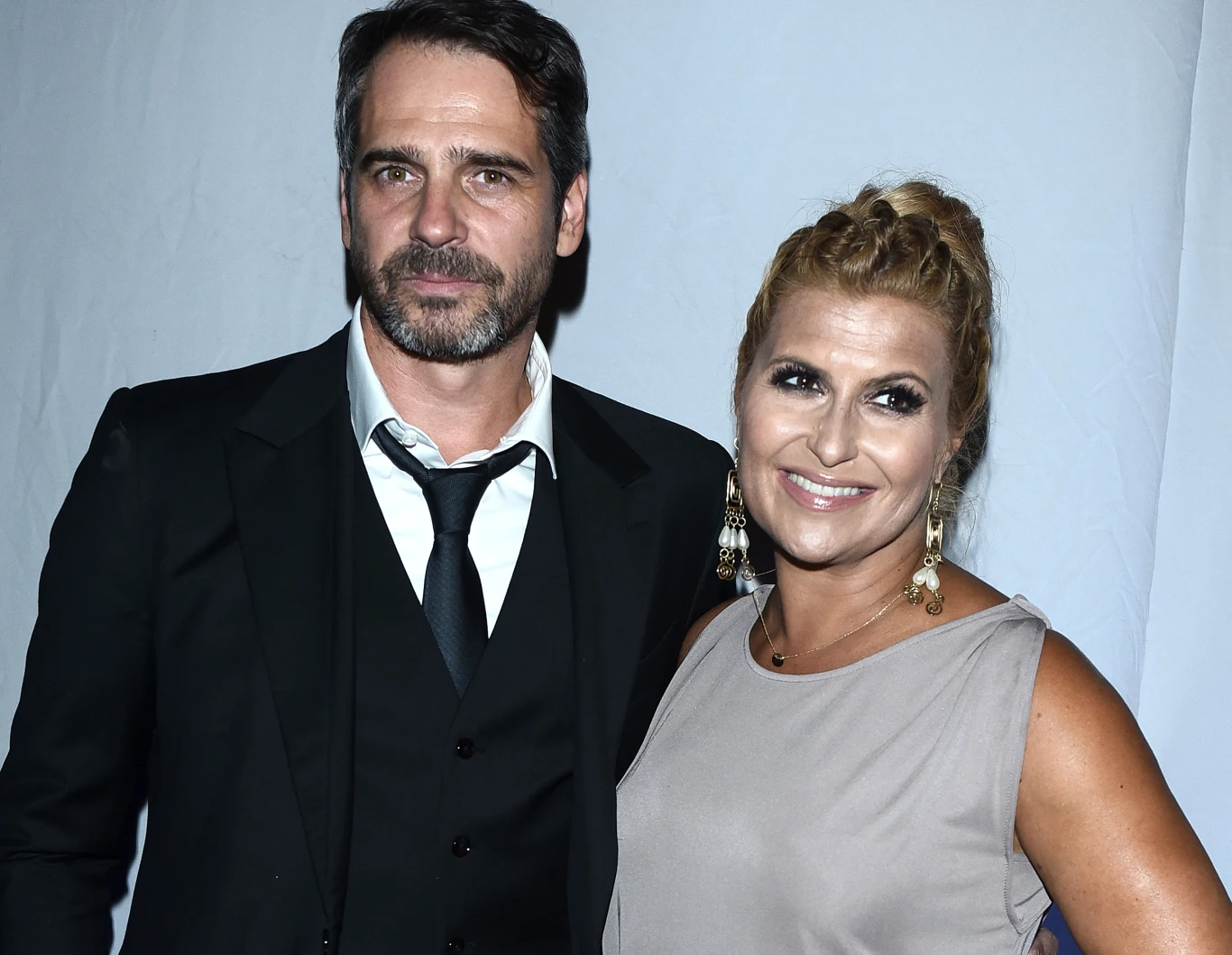 Para elegancko ubranych osób pozuje razem na tle jasnej ścianki, mężczyzna ma na sobie czarny garnitur z białą koszulą i krawatem, kobieta jest ubrana w jasną sukienkę, ma upięte włosy i ozdobne kolczyki, oboje patrzą w obiektyw i uśmiechają się.