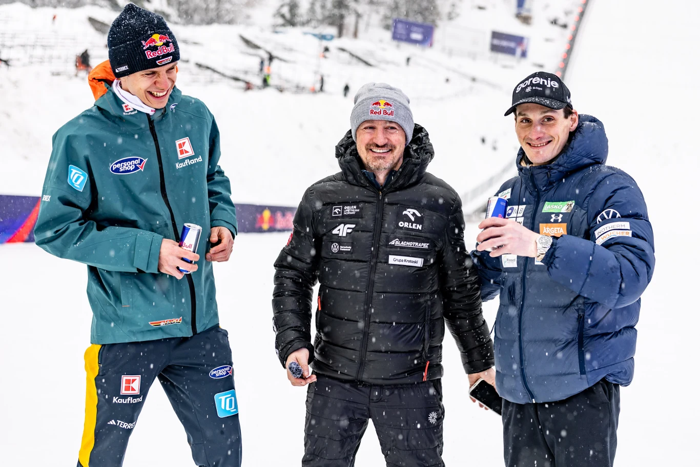 Trzech mężczyzn ubranych w sportowe kurtki zimowe i czapki stoi razem na śniegu, uśmiechając się i trzymając puszki napoju, w tle ośnieżona skocznia narciarska.