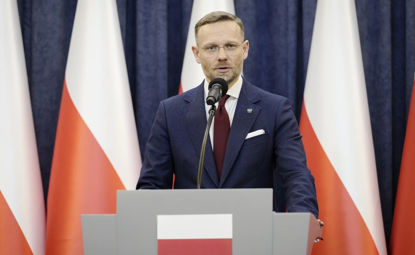 mężczyzna w garniturze i krawacie stoi za mównicą z godłem Polski, przemawiając do mikrofonu na tle kilku polskich flag