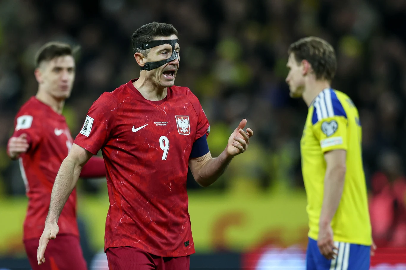 Piłkarz w czerwonym stroju z numerem 9, mający opaskę na twarzy, gestykuluje podczas meczu piłkarskiego; w tle inni zawodnicy w czerwonych i żółto-niebieskich strojach.