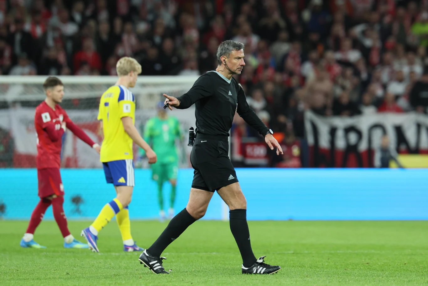 arbiter w czarnym stroju prowadzi mecz piłkarski na stadionie, w tle widoczni piłkarze w czerwonych i żółtych koszulkach oraz rozmyta widownia