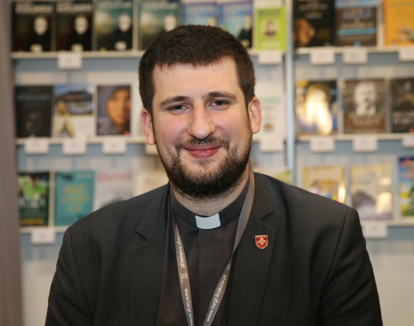 mężczyzna w koloratce i ciemnej marynarce z przypiętą odznaką, siedzi przed regałem z książkami, uśmiecha się patrząc wprost na obiektyw