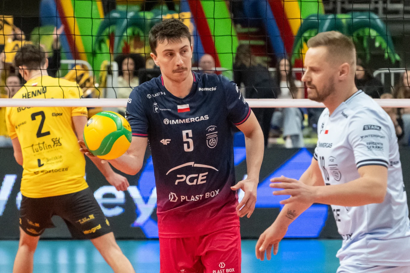 siatkarz w granatowej koszulce z numerem 5 trzyma piłkę przy siatce podczas meczu, obok niego siatkarz w białej koszulce, w tle zawodnicy drużyny w żółtych strojach