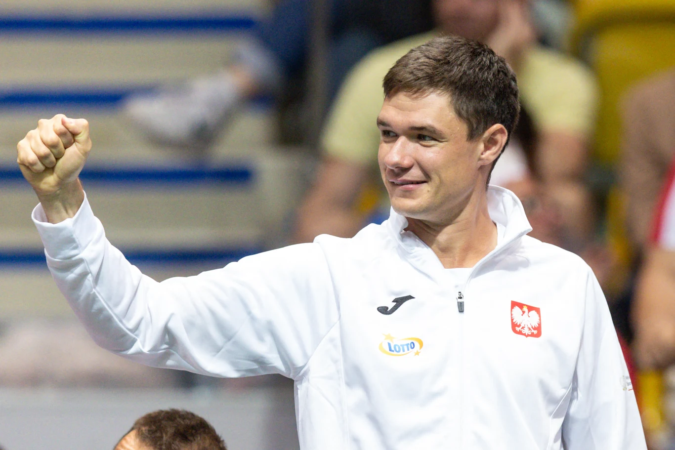 Mężczyzna w białej sportowej kurtce z godłem Polski oraz logotypami Lotto i Joma, zaciśnięta pięść uniesiona w geście triumfu, uśmiechnięta twarz, rozmyte tło z siedzącymi osobami.