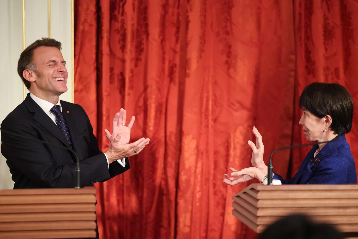 Dwoje polityków stoi przy mównicach na tle czerwonych zasłon, prowadząc rozmowę z dużą ekspresją — mężczyzna śmieje się z szeroko otwartymi dłońmi, a kobieta odpowiada gestami rękami.