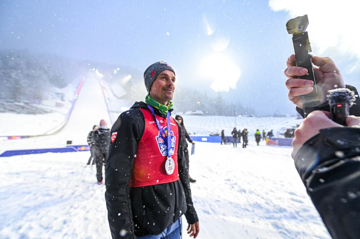 sportowiec w kurtce oraz czapce z medalem na szyi stoi naśnieżonym stadionie obok skoczni narciarskiej, w tle widoczni są dziennikarze i operatorzy nagrywający wydarzenie, a przed twarzą sportowca trzymana jest kamera przez inną osobę