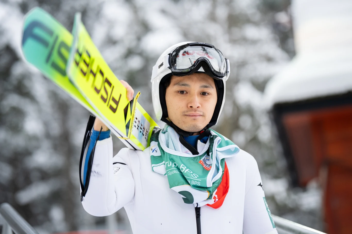 Skoczek narciarski w białym kombinezonie i kasku trzyma narty na ramieniu, na szyi zawieszony medal i zielony szalik z logo sponsora, w tle ośnieżone drzewa.