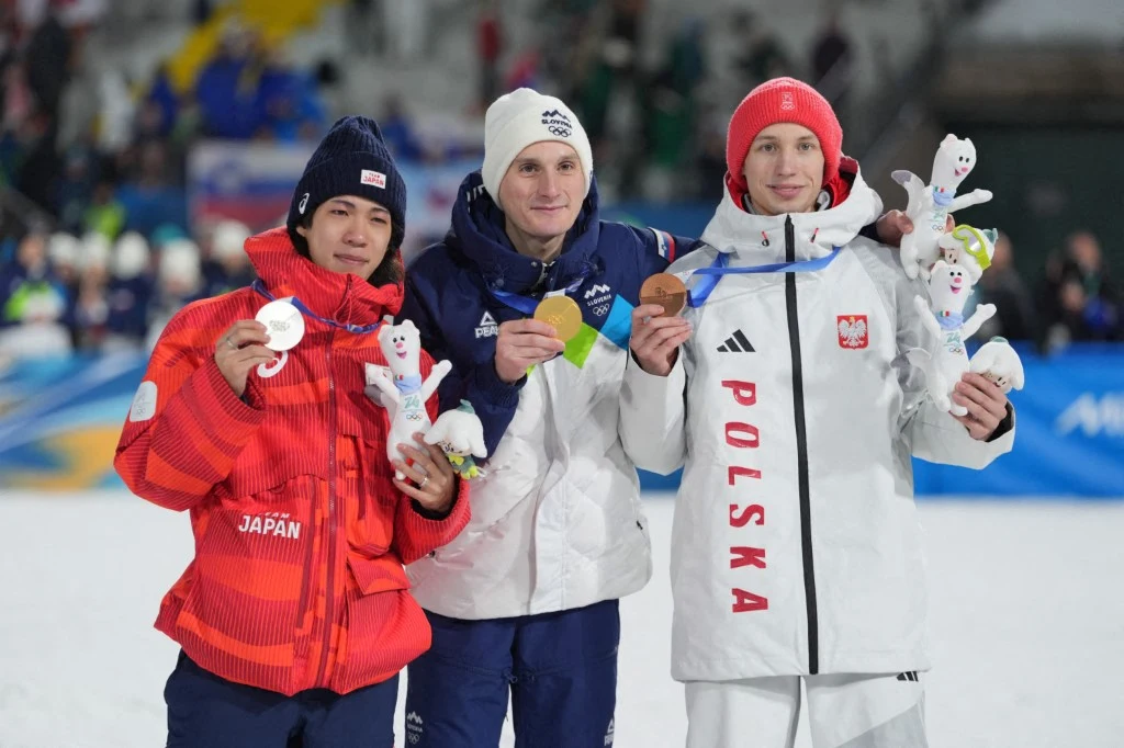 Trzech sportowców stojących na podium, każdy z medalem i maskotką, ubrani w stroje reprezentacyjne swoich krajów: Japonii, Finlandii i Polski, na tle zimowej areny sportowej.