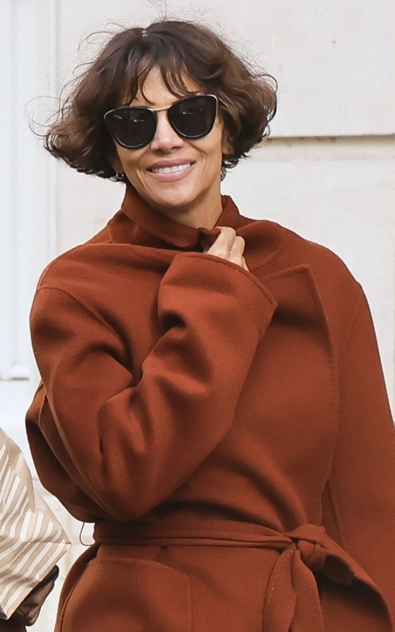 kobieta z krótkimi, ciemnymi włosami i okularami przeciwsłonecznymi uśmiecha się, ubrana w długi, brązowy płaszcz wiązany w pasie