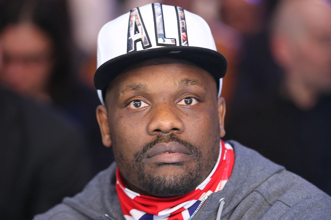 mężczyzna w białej czapce z napisem 'ALI', ubrany w szarą bluzę i czerwoną chustę z motywem flagi, patrzy przed siebie z poważnym wyrazem twarzy