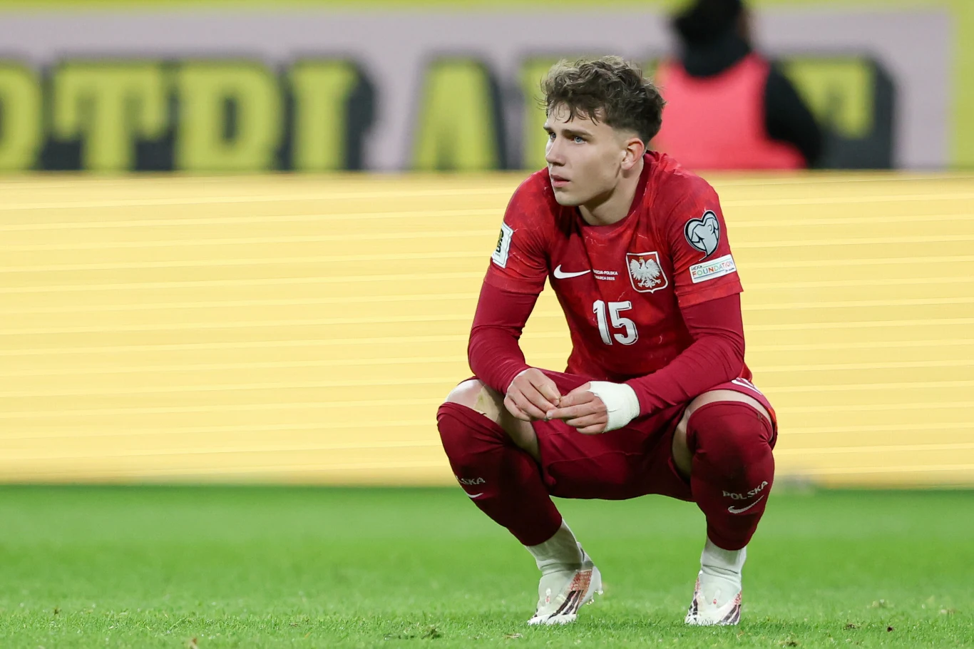 piłkarz w czerwonym stroju z numerem 15 i godłem Polski na piersi kuca na murawie stadionu z wyraźnie zamyśloną miną, w tle widoczna rozmyta publiczność oraz żółta reklama