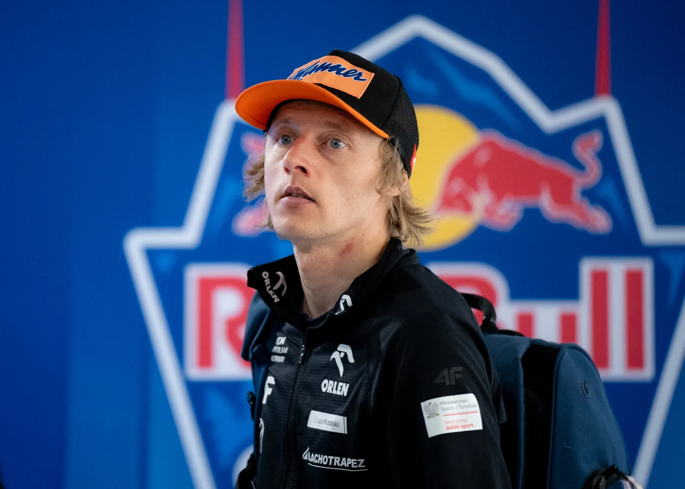 Młody mężczyzna w czarnej kurtce i czapce z daszkiem na tle dużego logo Red Bull, z plecakiem na ramieniu, patrzy lekko w górę.