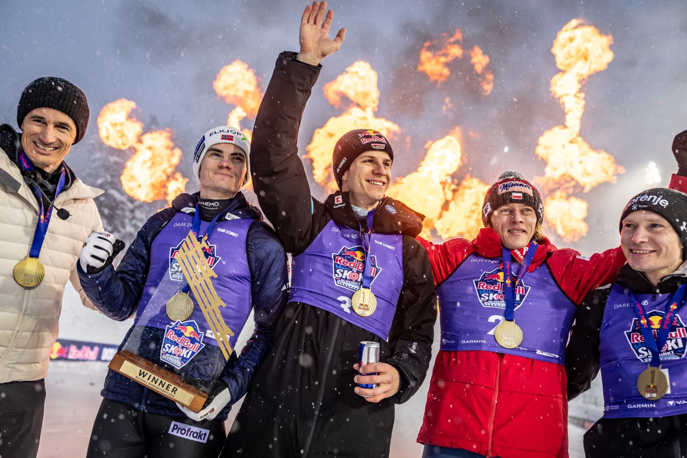 Grupa pięciu uśmiechniętych sportowców ubranych w zimowe stroje, każdy z medalem na szyi, jeden trzyma trofeum, w tle spektakularne wybuchy ognia na śnieżnym tle, co podkreśla atmosferę triumfu i sukcesu.