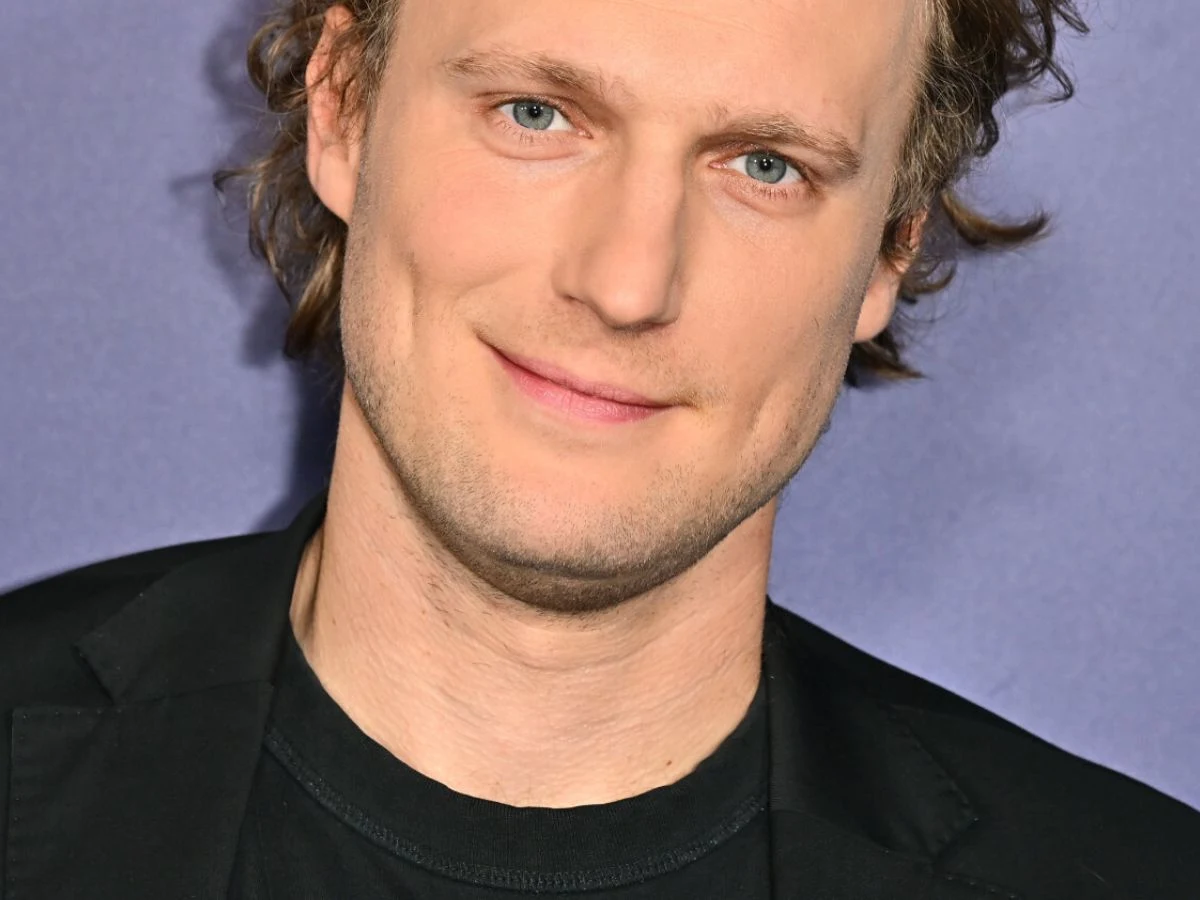 Uśmiechnięty mężczyzna z jasną cerą i niebieskimi oczami, ubrany w czarny t-shirt oraz czarną marynarkę, na fioletowym tle.