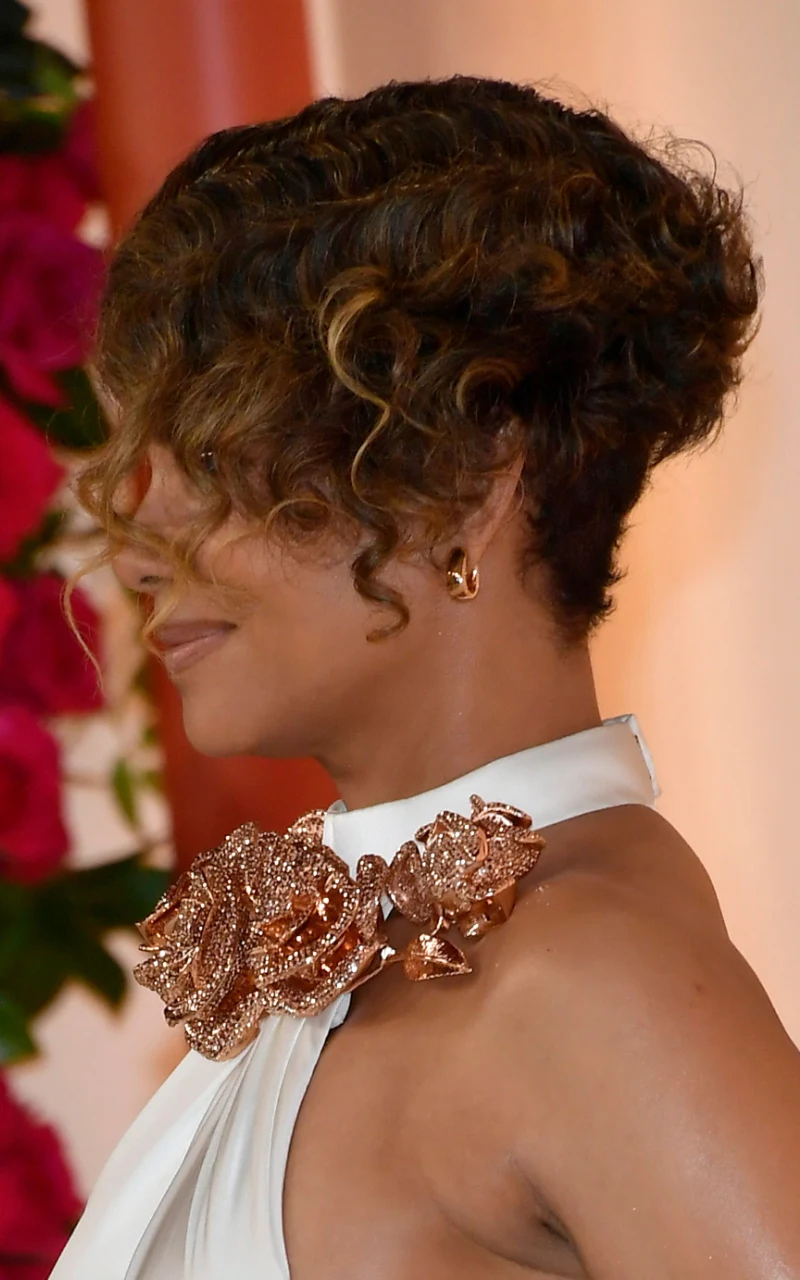 Kobieta z krótkimi, kręconymi włosami w eleganckiej fryzurze, ubrana w biały strój ozdobiony dużą, efektowną broszką w kształcie kwiatów w złotym kolorze, na tle dekoracji z różowych kwiatów.