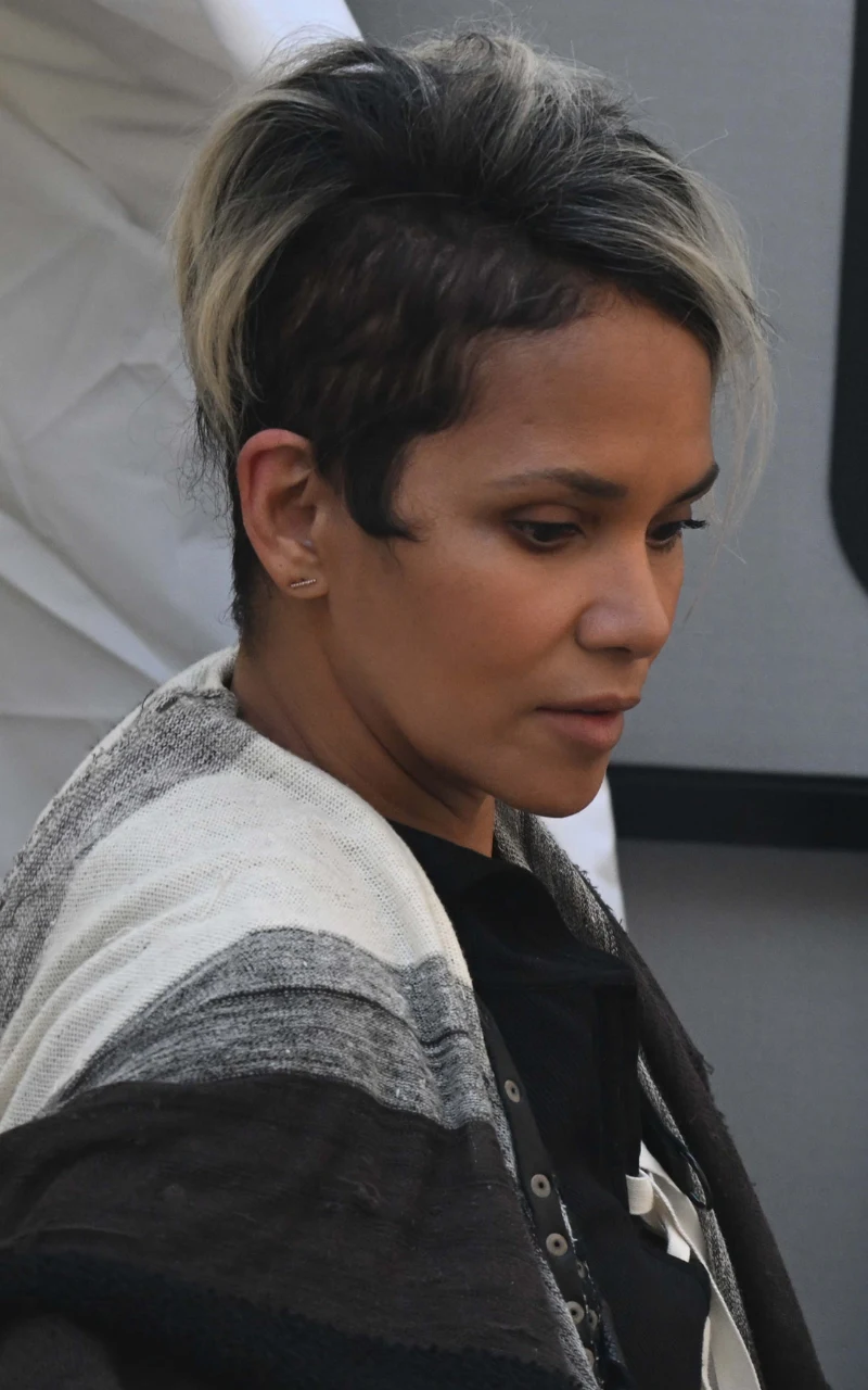 Kobieta o ciemnej karnacji i krótkich, ciemnych włosach z jasnymi pasmami, ubrana w czarno-białą narzutkę, patrzy zamyślona w dół, otoczona neutralnym tłem.