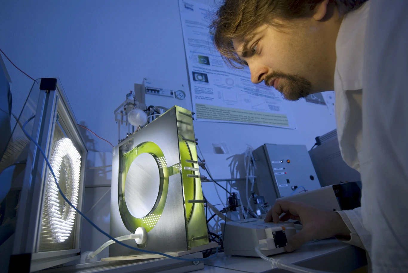 mężczyzna w białym fartuchu laboratoryjnym obserwuje skomplikowane urządzenie z podświetlanym okrągłym elementem, wokół którego rozmieszczone są liczne przewody oraz aparatura naukowa