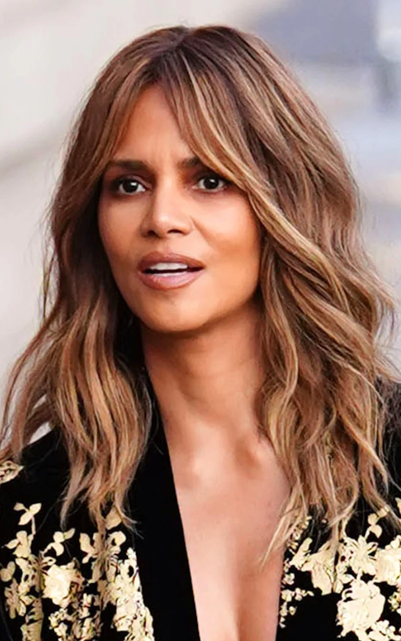 Kobieta o długich falowanych włosach w odcieniu jasnobrązowym, ubrana w czarną marynarkę ozdobioną złotymi haftami, patrzy przed siebie z lekkim uśmiechem na twarzy.