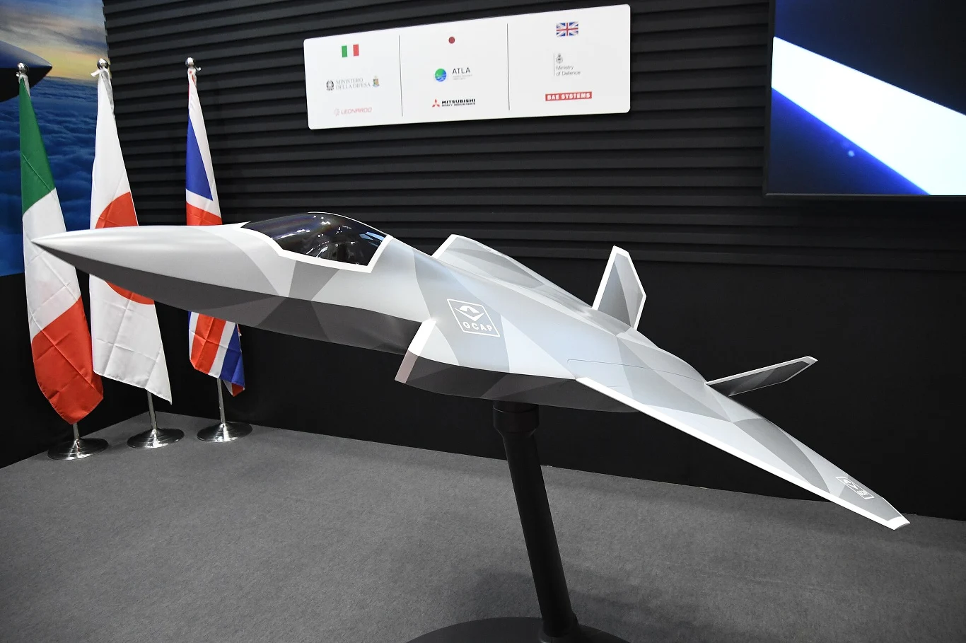 makieta futurystycznego myśliwca odrzutowego o kanciastych, aerodynamicznych kształtach, ustawiona na czarnym stojaku, w tle trzy flagi narodowe: Włoch, Japonii i Wielkiej Brytanii oraz tablica informacyjna z logotypami firm i narodowymi flagami