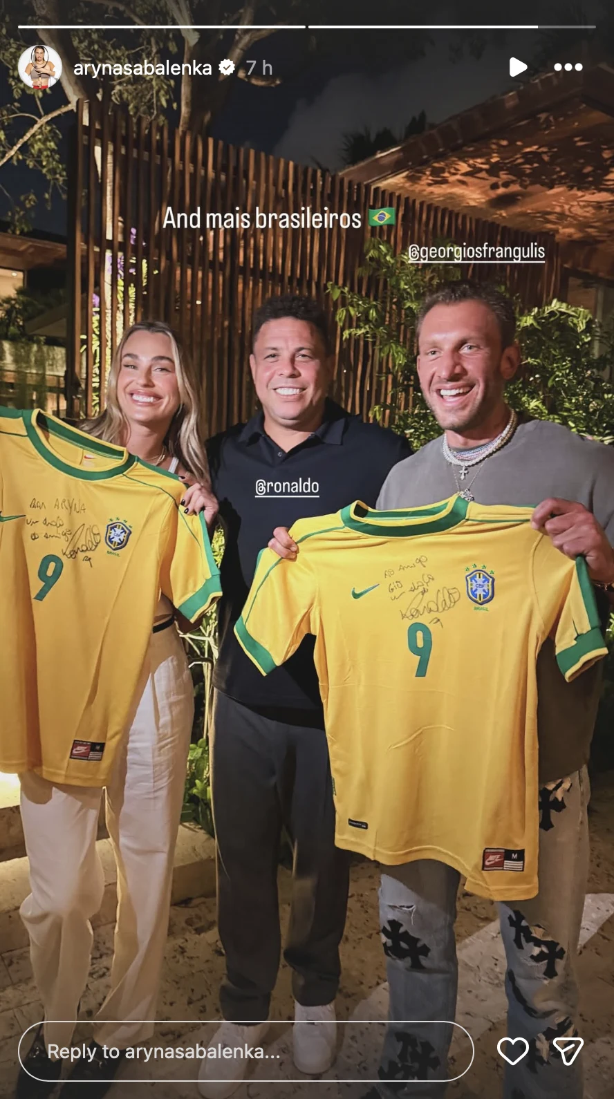 Trzy uśmiechnięte osoby stoją razem na tle roślin i drewnianych konstrukcji, dwie z nich trzymają żółte koszulki reprezentacji Brazylii z autografami i numerem 9, środkowa osoba ubrana jest na czarno.