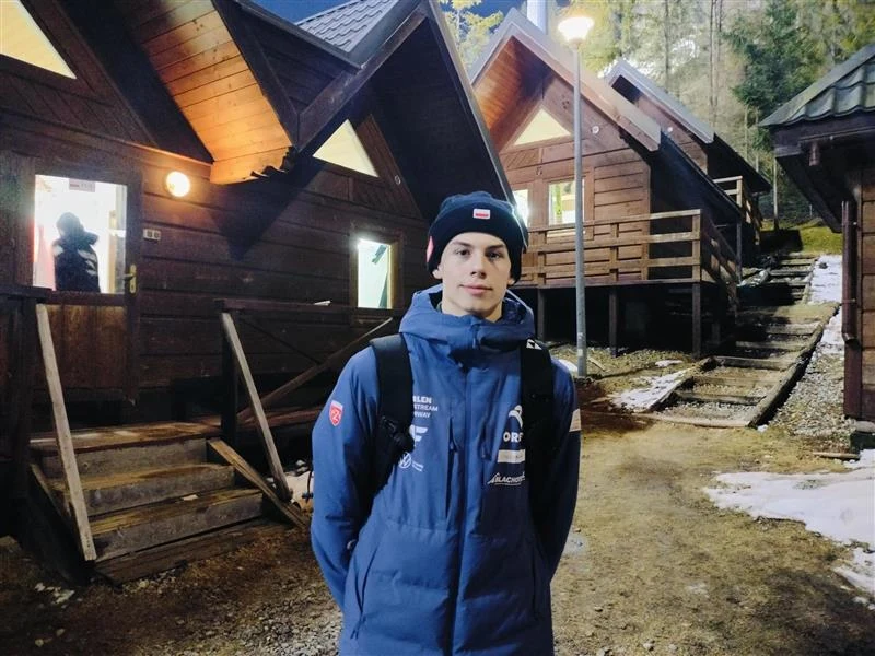 młody mężczyzna w sportowej kurtce i czapce stoi przed drewnianymi domkami górskimi, w tle wieczorna sceneria z zapalonymi światłami oraz resztkami śniegu na ziemi