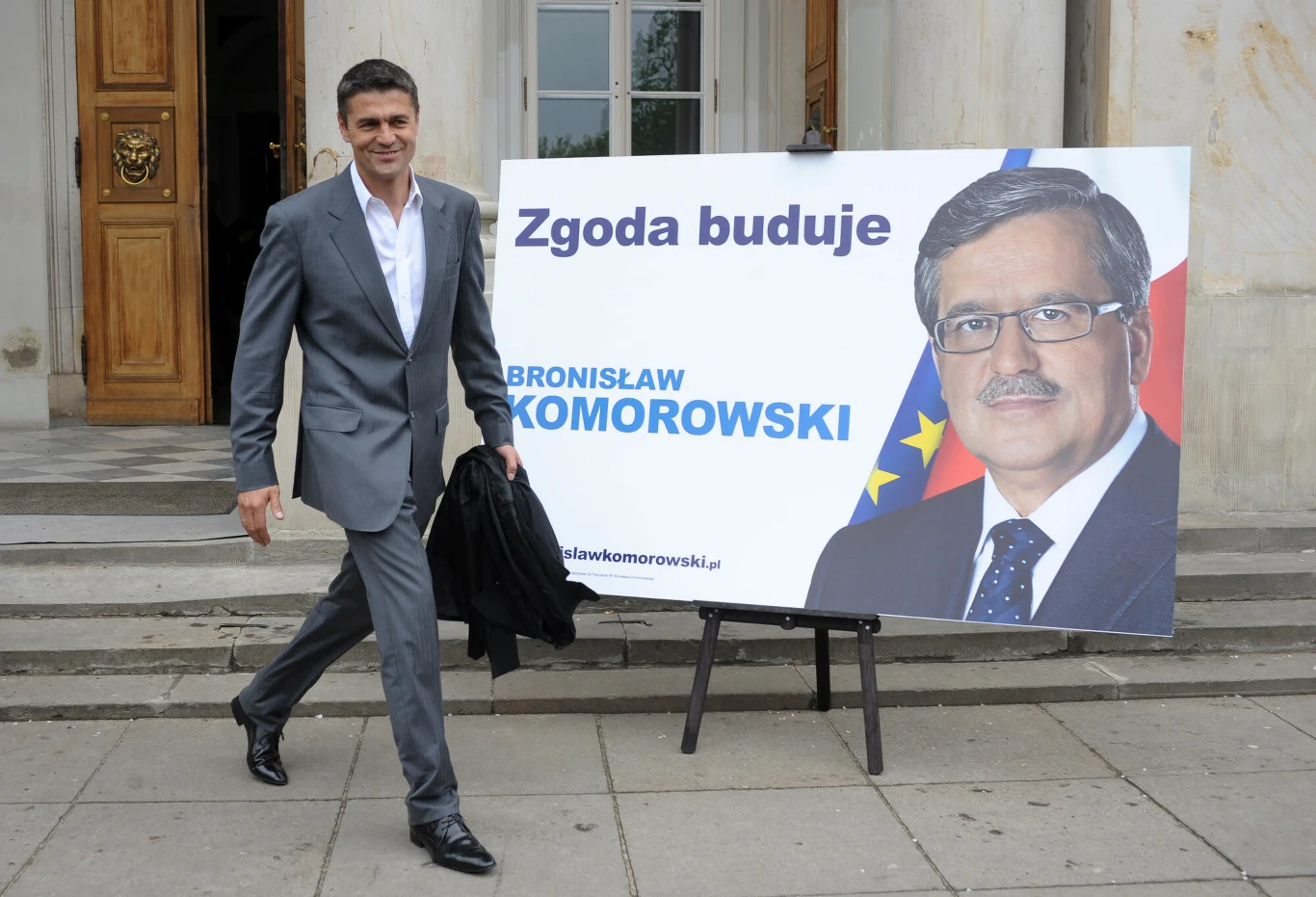mężczyzna w szarym garniturze idzie chodnikiem obok dużego billboardu wyborczego z portretem Bronisława Komorowskiego, hasłem „Zgoda buduje” oraz flagami w tle