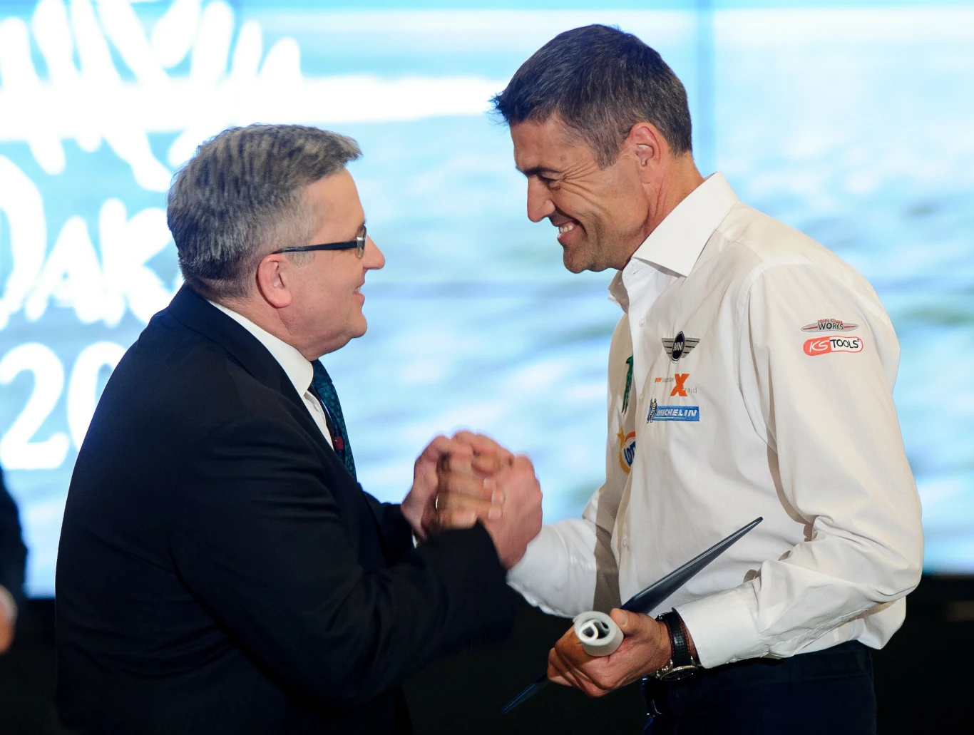 Dwóch mężczyzn uśmiecha się do siebie podczas serdecznego uścisku dłoni; jeden z nich ubrany w garnitur, drugi w białą koszulę z naszywkami, trzymając w ręku dokument oraz zwiniętą kartkę.