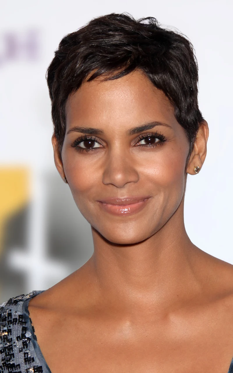 kobieta o krótkich, ciemnych włosach i brązowych oczach z delikatnym makijażem, uśmiechnięta, ubrana w elegancką, błyszczącą suknię na jasnym tle