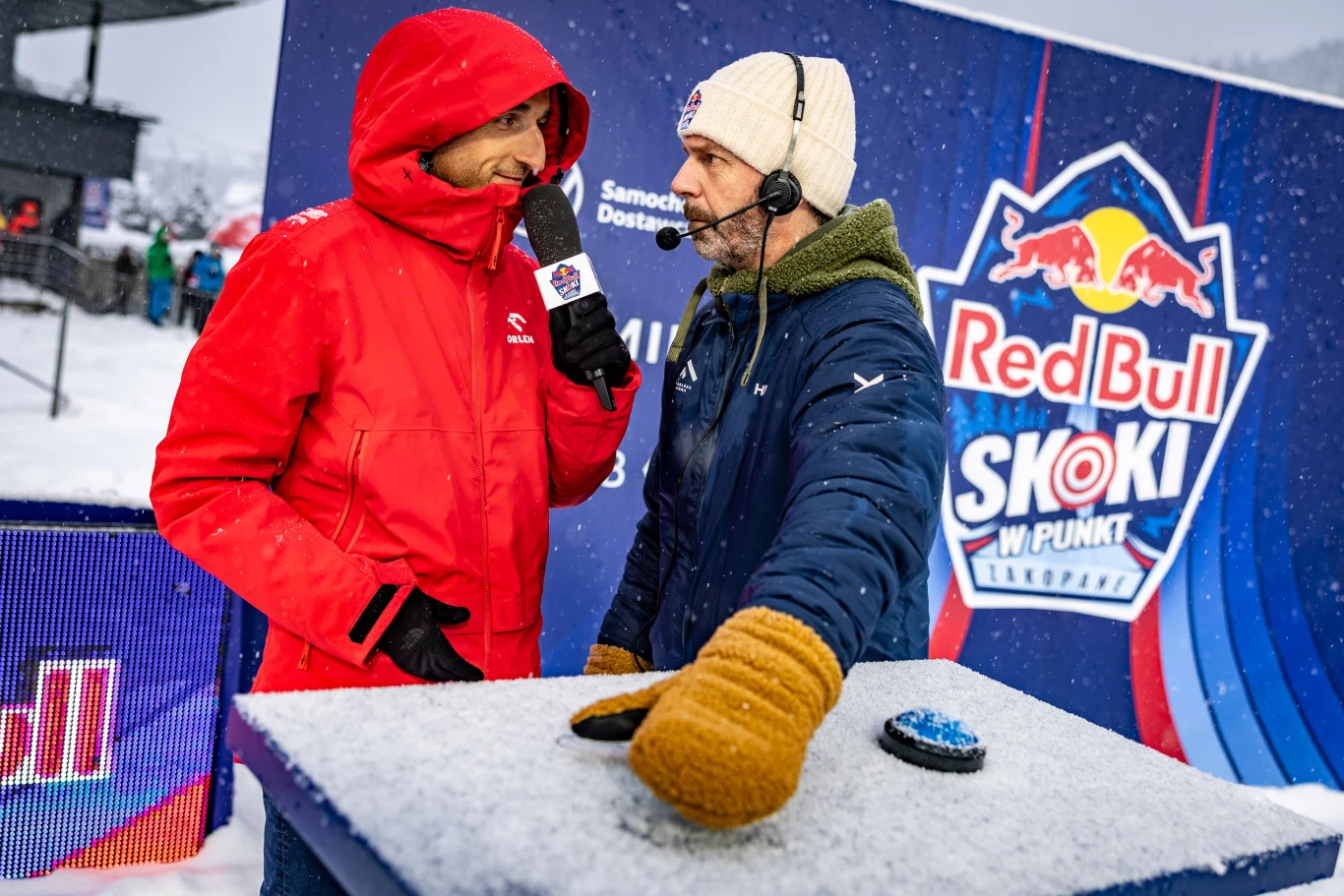 Dwóch mężczyzn rozmawia podczas zimowej imprezy sportowej; jeden ma na sobie czerwoną kurtkę i trzyma mikrofon, drugi nosi granatową kurtkę i beżową czapkę, z dużymi żółtymi rękawicami; między nimi znajduje się przycisk na zamarzniętym stole, a w tle w...