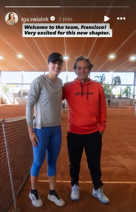 dwie osoby stoją razem na krytym korcie tenisowym, jedna z nich ubrana w sportową odzież oraz czapkę z daszkiem, druga w czerwonej bluzie i czarnych spodniach, obie uśmiechnięte, w tle widoczne palmy i przeszklone ściany