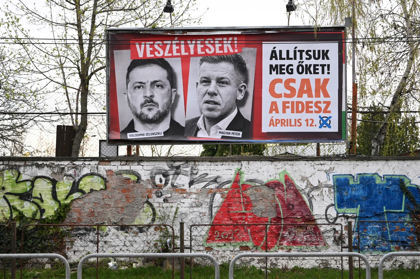 duży plakat wyborczy na tle ogrodzenia pokrytego kolorowym graffiti, z wizerunkami Wołodymyra Zełenskiego i Roberta Fica oraz wyraźnymi napisami w języku węgierskim, nawołującymi do głosowania przeciwko nim i poparcia partii Fidesz, obok widoczne drzew...