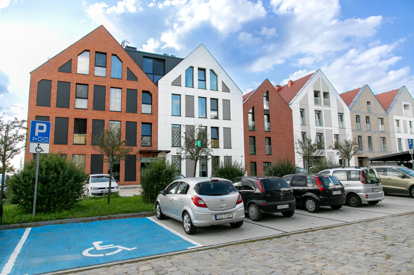 Dopłata do mieszkania dla osoby niepełnosprawnej. Ile pieniędzy można otrzymać? Nowoczesne budynki mieszkalne z cegły i białą elewacją, parking z miejscem dla osób niepełnosprawnych.