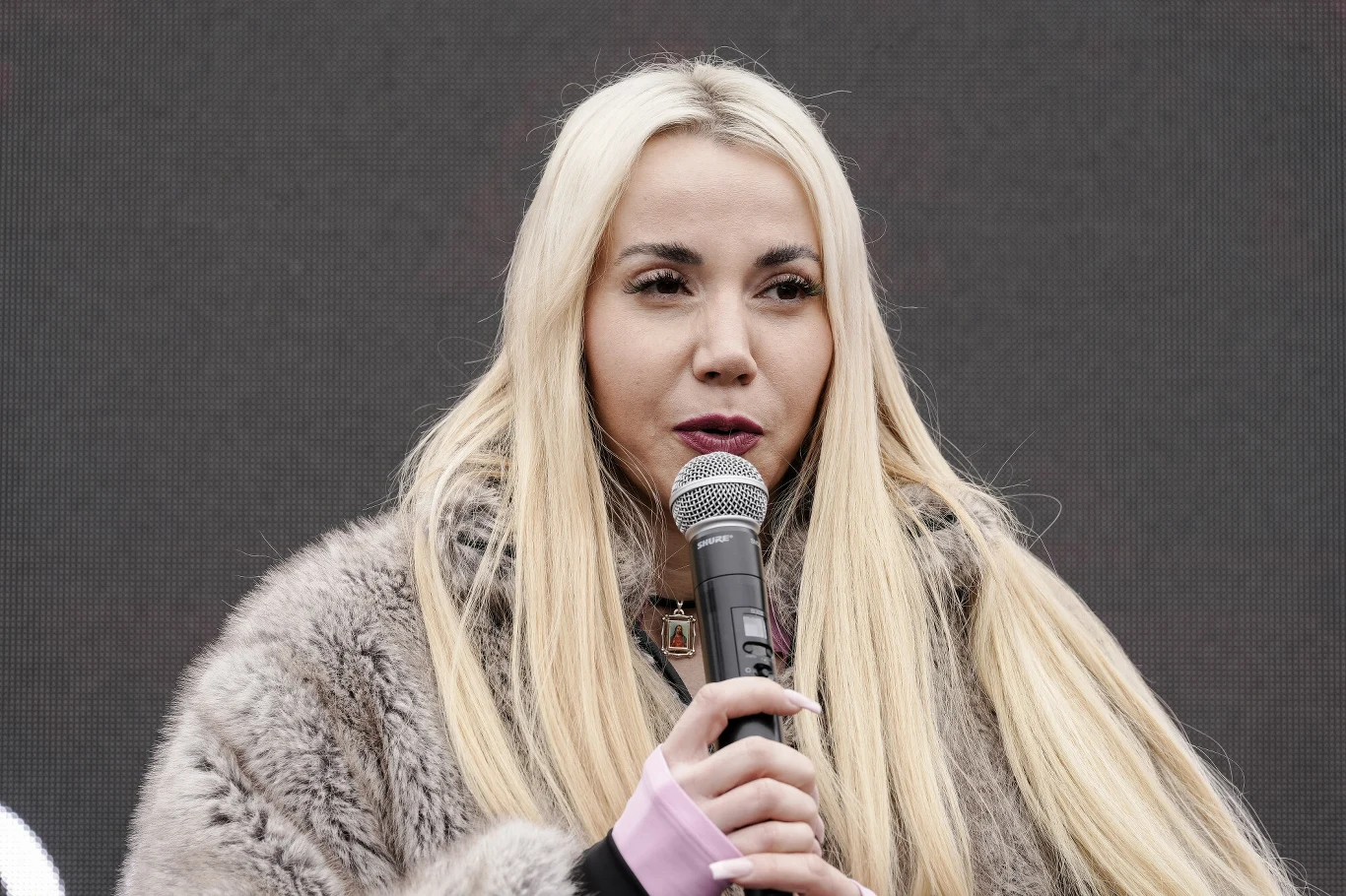 Marianna Schreiber Kobieta z długimi blond włosami trzyma mikrofon i przemawia, ubrana w szarą futrzaną kurtkę, na tle jednolitej ciemnej powierzchni.