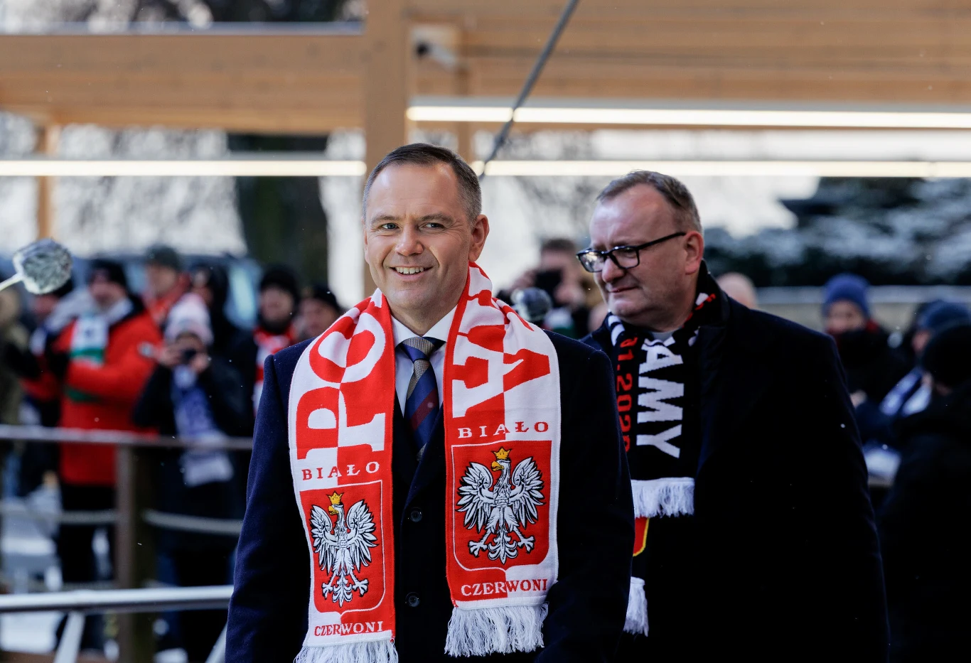 dwóch mężczyzn ubranych w eleganckie garnitury i biało-czerwone szaliki z orłem oraz napisami patriotycznymi, stojących na tle grupy ludzi w zimowej scenerii