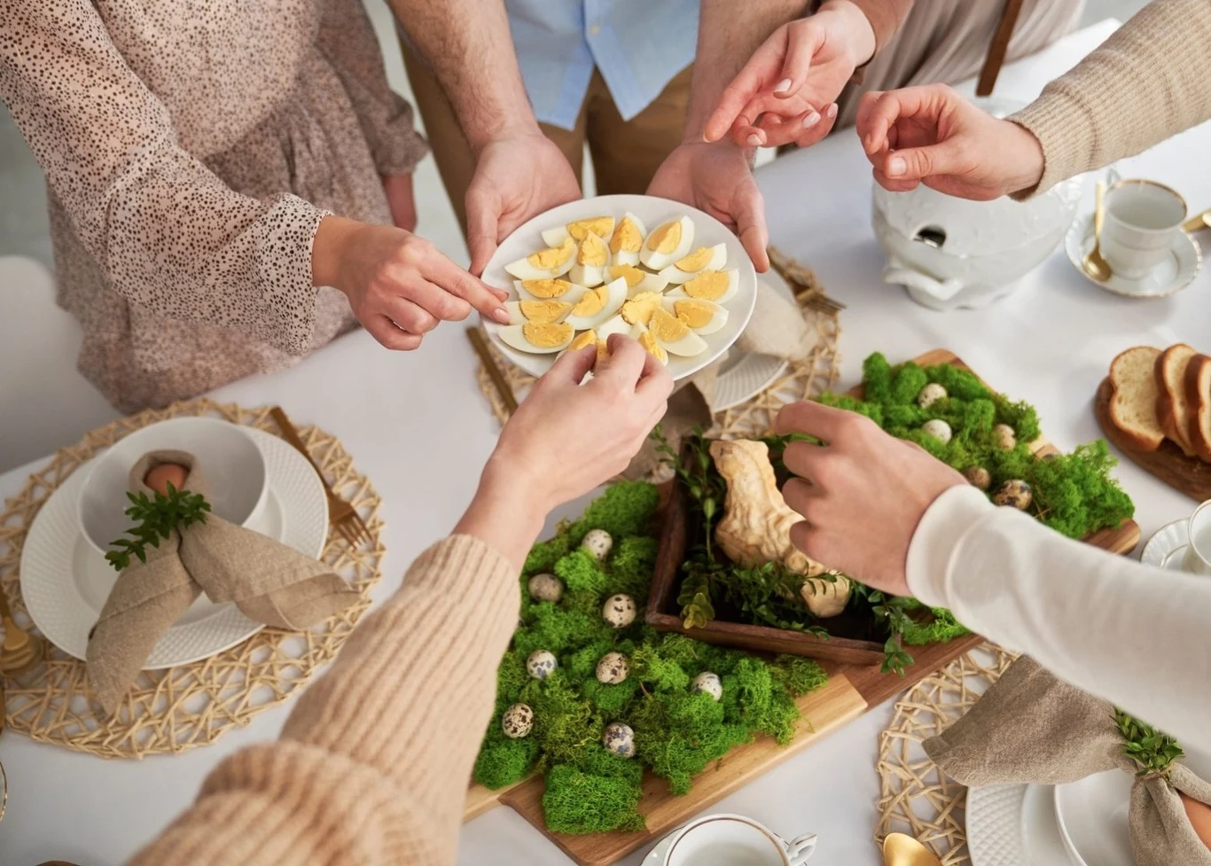 Kilka osób sięga po jajka na półmisku, na świątecznym stole udekorowanym zielonym mchem, przepiórczymi jajkami i pieczywem, wokół elegancko nakryte talerze z lnianymi serwetkami.