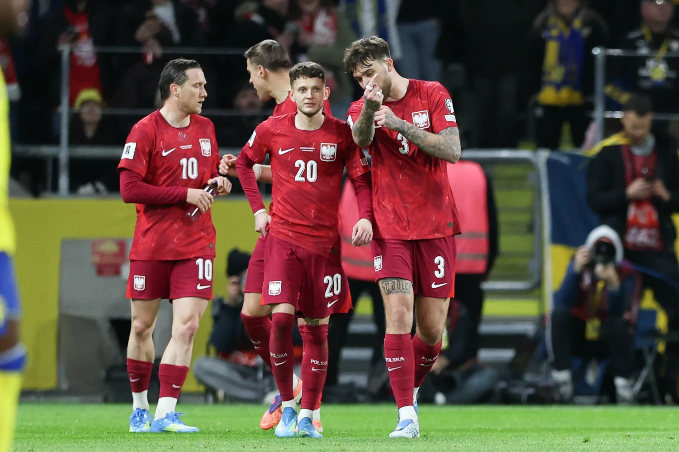 czterech polskich piłkarzy w czerwonych strojach na murawie, skupionych na rozmowie, otoczonych przez żółto-niebiesko ubranych rywali oraz kibiców na trybunach