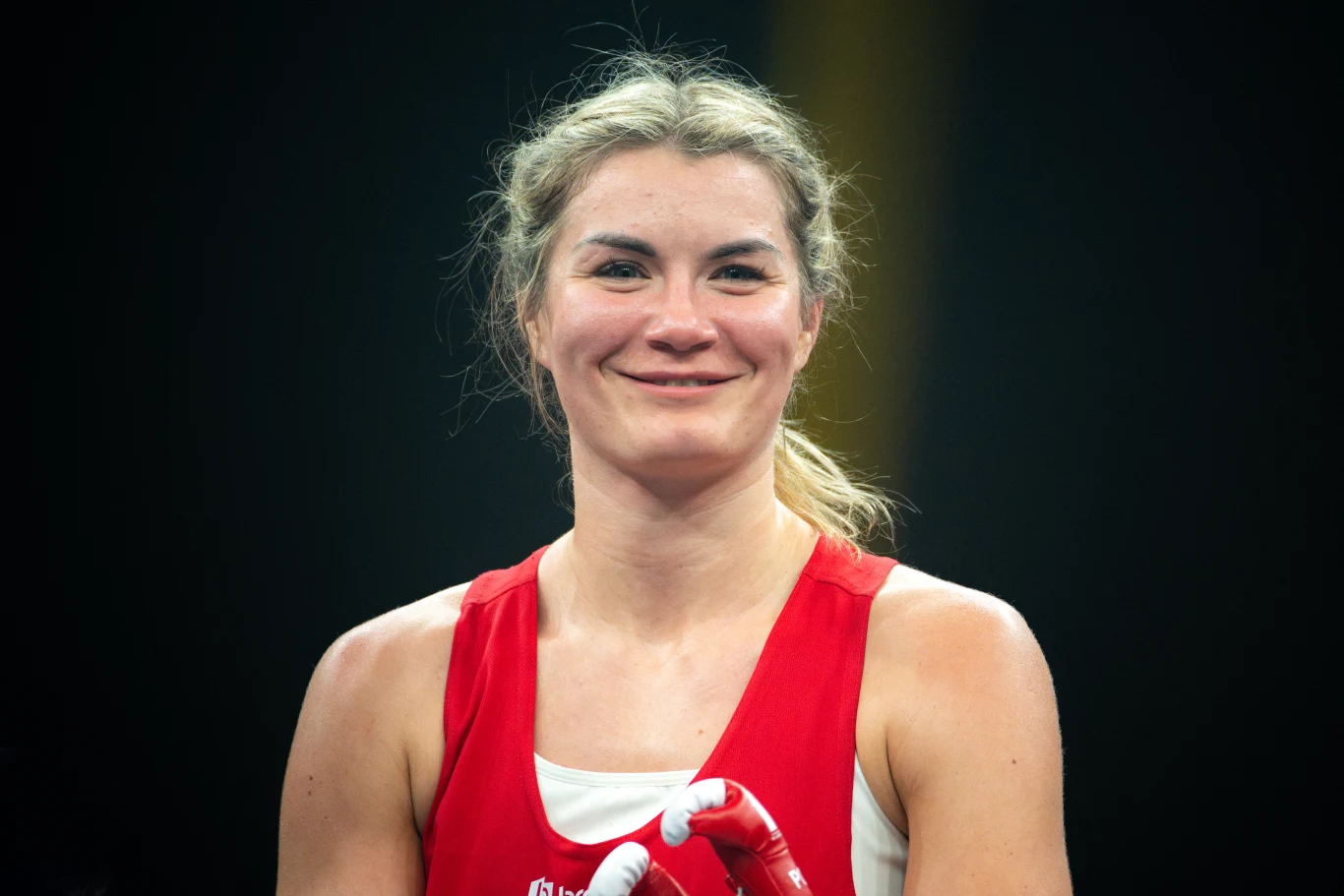 uśmiechnięta kobieta w sportowym ubraniu i rękawicach bokserskich stoi na tle ciemnego, rozmytego tła