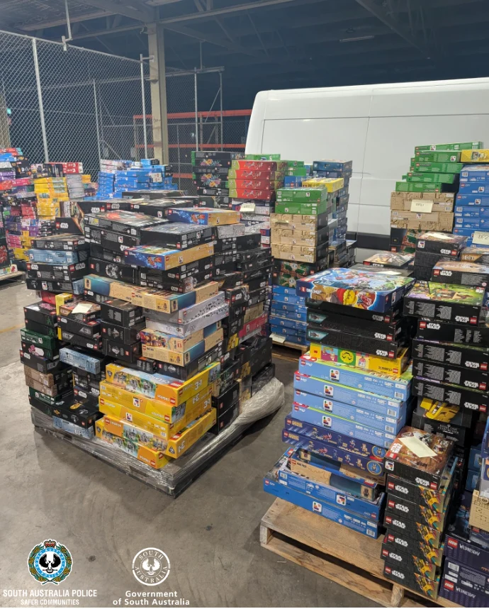 Stosy kolorowych pudełek z zestawami klocków LEGO ustawione na paletach w magazynie, w tle biały samochód dostawczy i metalowe ogrodzenie, na dole logosy South Australia Police i Government of South Australia.