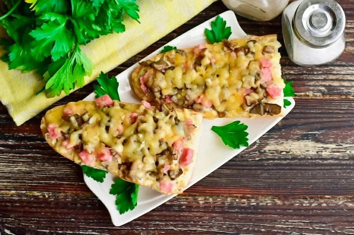 Zapiekanki z pieczarkami i szynką wychodzą przyjemnie chrupiące Dwie zapiekanki z pieczarkami, szynką i serem na białym talerzu, z natką pietruszki obok soli i pieprzu.