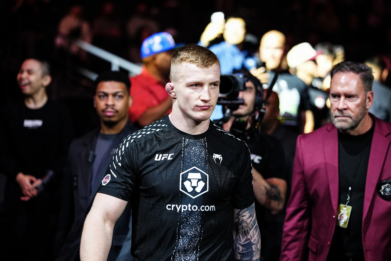 zawodnik UFC w czarnej koszulce z logo sponsora wychodzi na arenę otoczony przez ochroniarzy i ludzi z obsługi, w tle tłum kibiców
