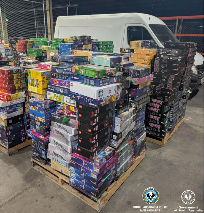 Kilka stosów pudełek z klockami LEGO ustawionych na paletach w magazynie, obok biały van, w tle metalowy płot i fragment hali magazynowej; na pierwszym planie logotypy południowo-australijskiej policji oraz rządu Australii Południowej.