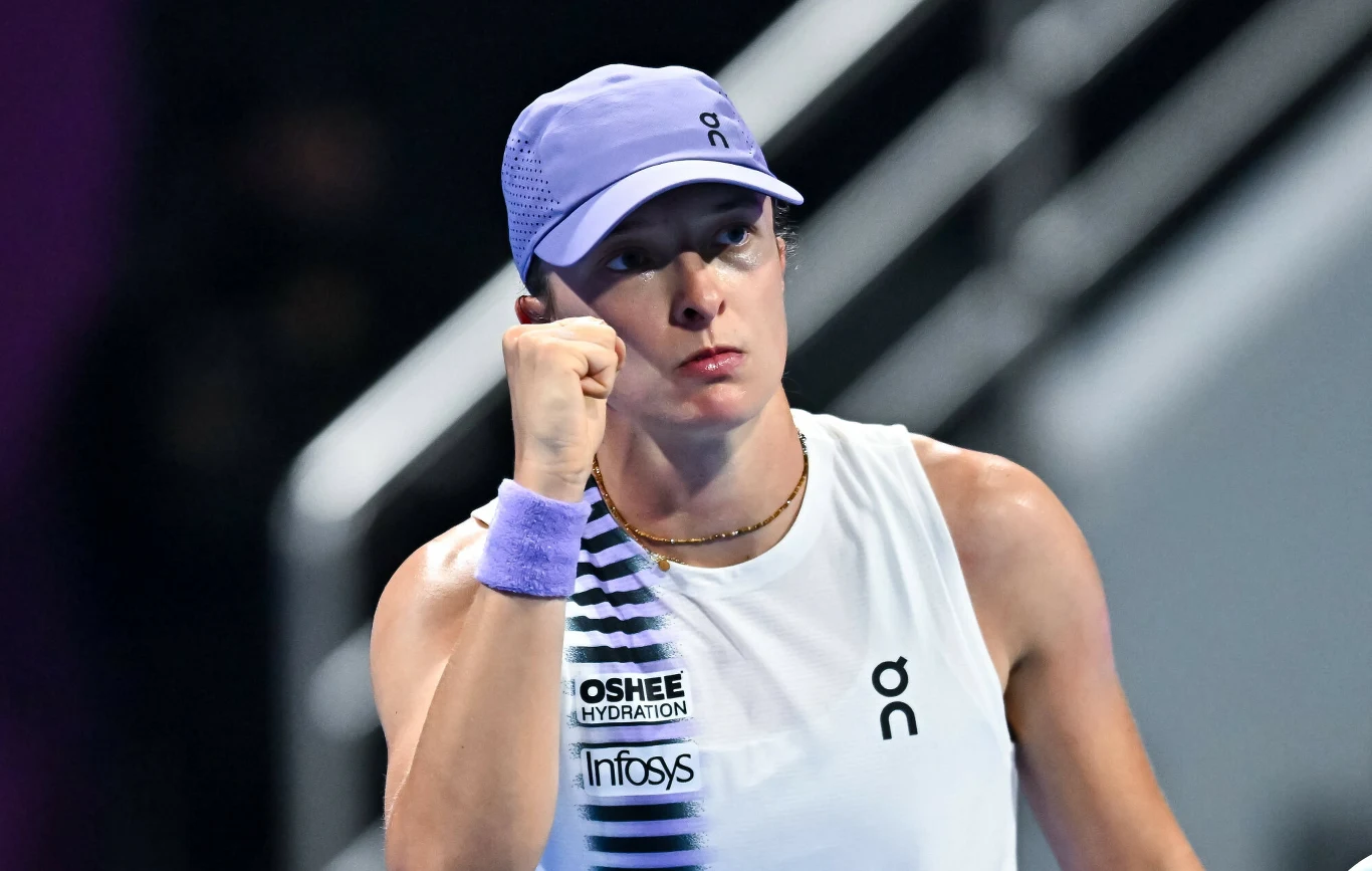 Iga Świątek Tenisistka ubrana w białą koszulkę sportową i fioletową czapkę z daszkiem zaciska pięść w geście determinacji podczas meczu na korcie tenisowym, z wyrazem skupienia na twarzy.