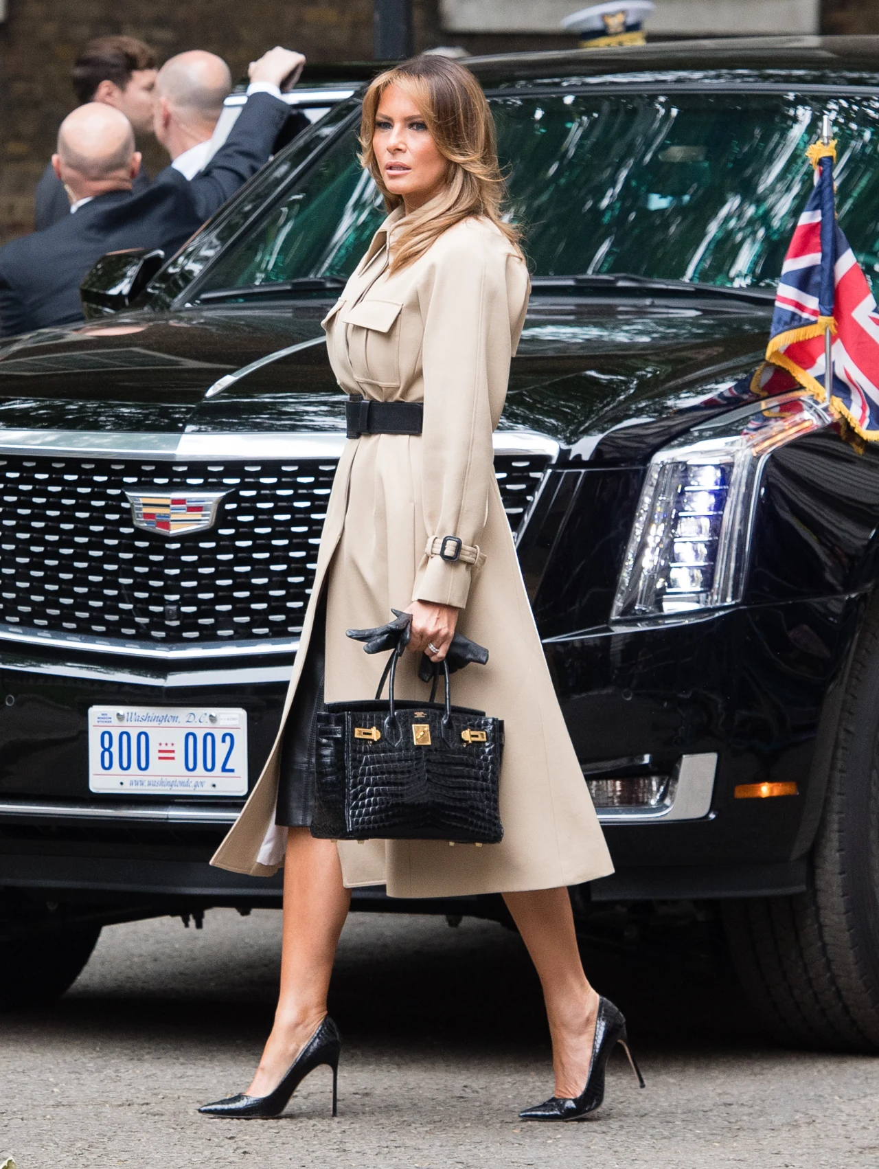 elegancka kobieta w beżowym trenczu i czarnych szpilkach przechodzi obok luksusowego, czarnego samochodu z widoczną flagą Stanów Zjednoczonych, trzymając czarną torebkę w ręce