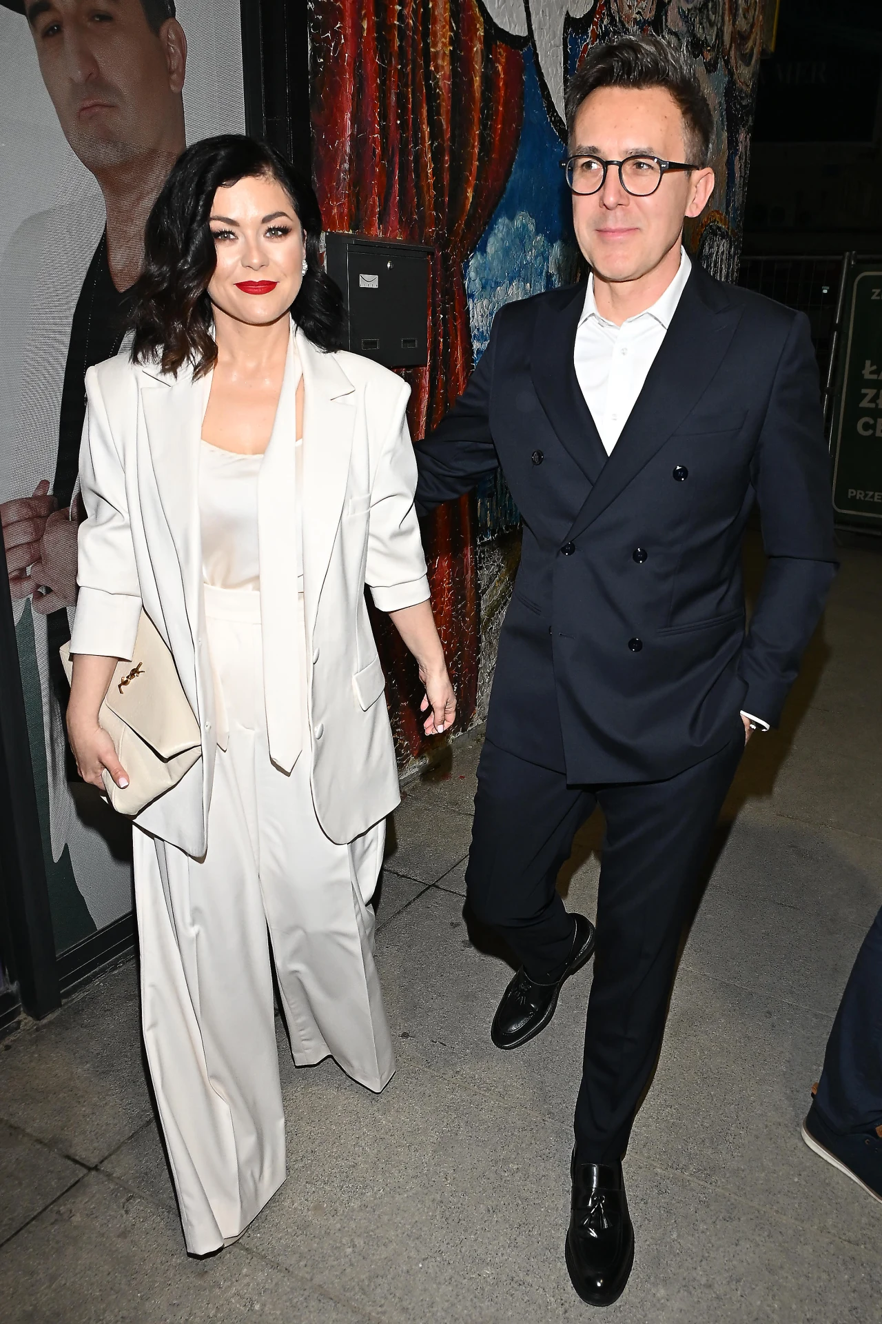 kobieta w eleganckim jasnym garniturze i mężczyzna w ciemnym garniturze idą razem chodnikiem, trzymając się za ręce, w tle kolorowy mural oraz fragment plakatu z wizerunkiem mężczyzny